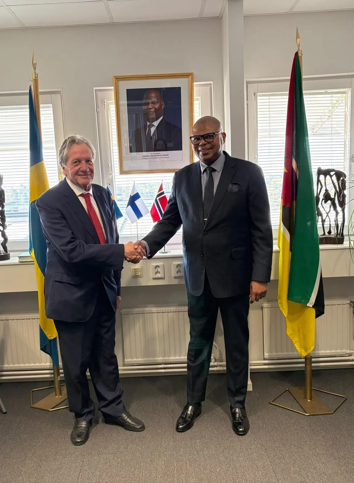 O Dr. Henric Thörnberg e o Embaixador Francisco Novela apertam as mãos na Embaixada em Estocolmo, diante do retrato do Presidente da República e entre as bandeiras da Suécia e de Moçambique