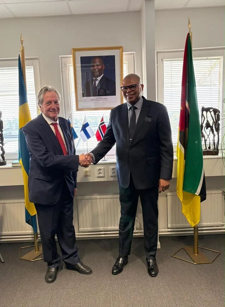 O Dr. Henric Thörnberg e o Embaixador Francisco Novela apertam as mãos na Embaixada em Estocolmo, diante do retrato do Presidente da República e entre as bandeiras da Suécia e de Moçambique