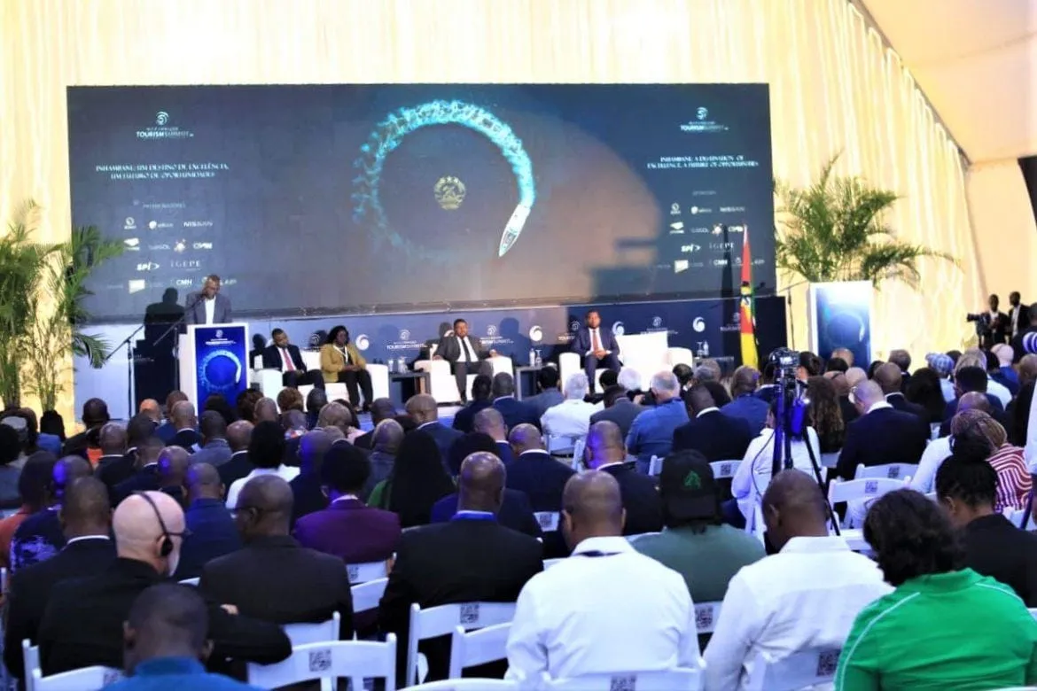 Sala de conferências do Mozambique Tourism Summit, com um orador ao púlpito e um painel sentado no palco, sob um grande ecrã com o logótipo do evento, e o público em primeiro plano